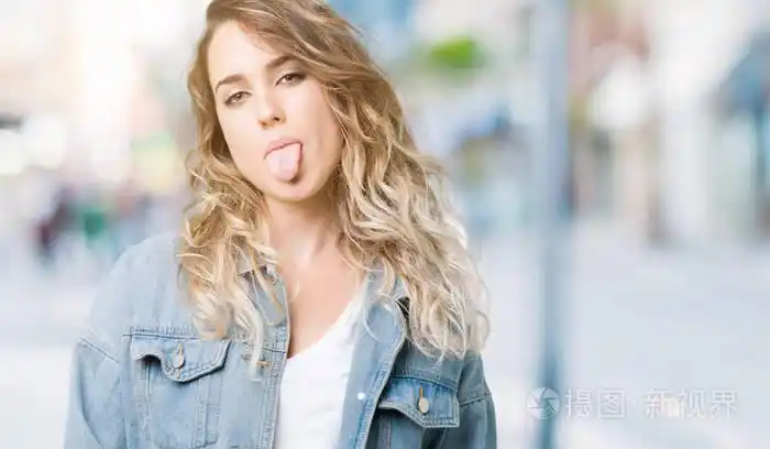 美丽的年轻金发女人穿着牛仔夹克在孤立的背景上伸出舌头,以有趣的