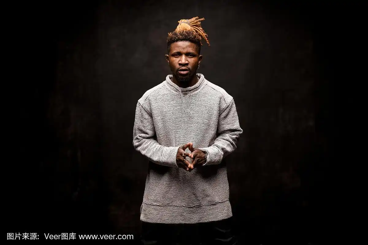 一名留着金色长发绺的黑人男子,手指紧握在一起,对着镜头摆姿势