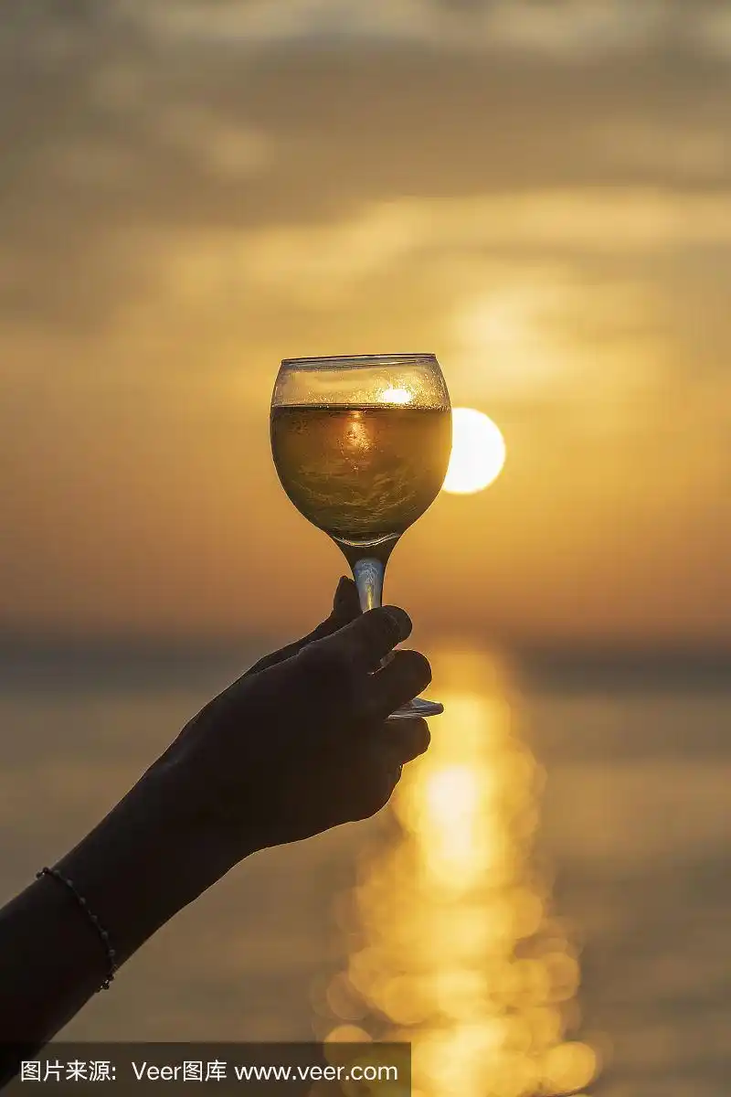 在热带海滩上,在美丽的日落下,手持酒杯的女人靠近海边.