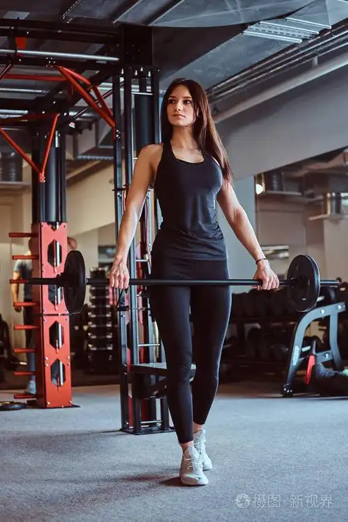 在健身俱乐部或健身房训练时, 漂亮的黑发女性在运动衫上持有杠铃照片