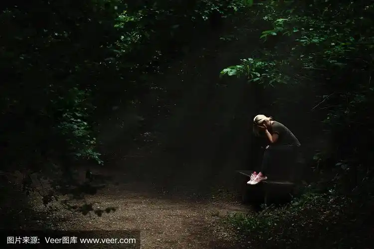 孤独而悲伤的金发女人坐在黑暗的森林小路中间的一个光点上的长椅上